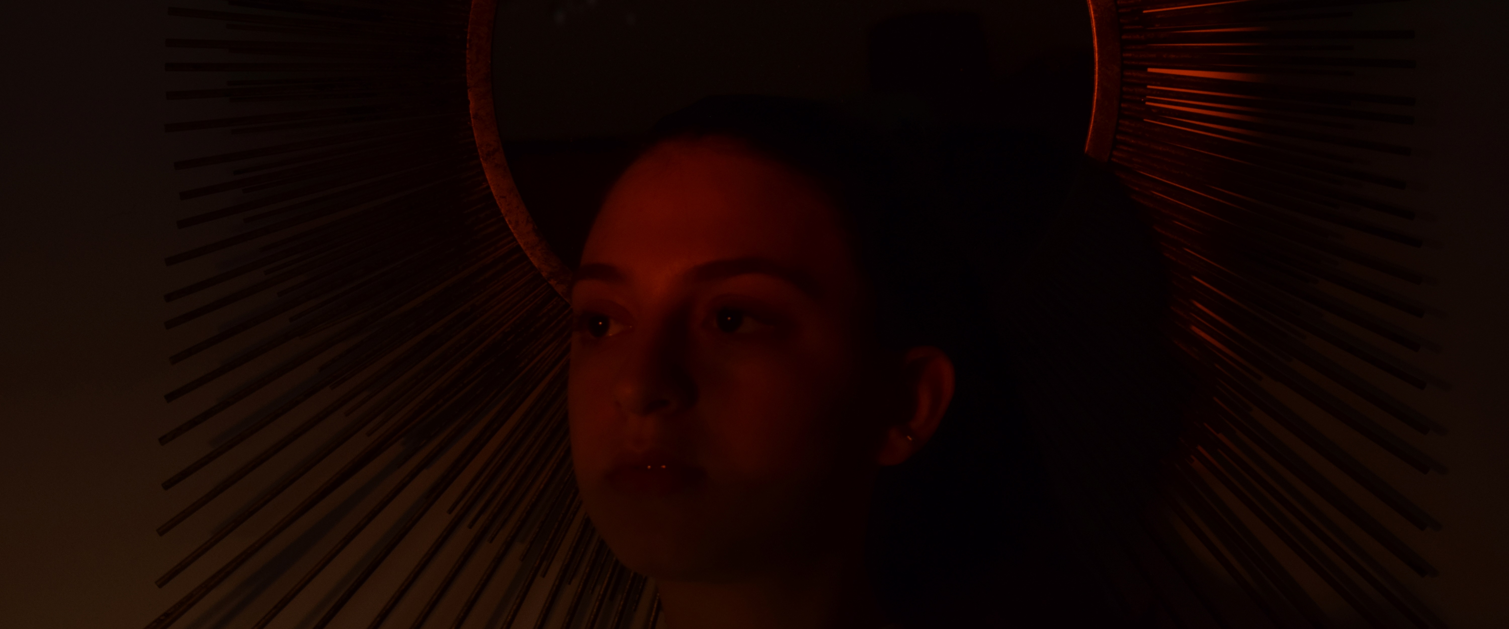 An image of a lady with a red hue from the setting sun, infront of a circle wall decoration surrounded by spikes which resembles the sun. shot by TayStudios.